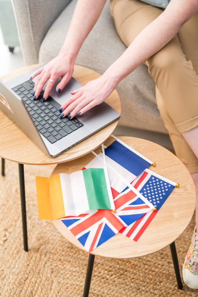 Une traductrice SEO travaille sur son ordinateur. Des drapeaux sont posés à côté sur une table basse : France, Irlande, USA, UK.
